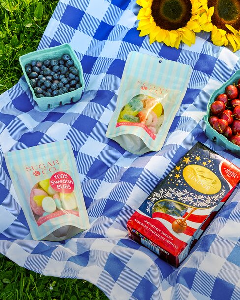 Picnic blanket styled with blueberries, cherries, assorted Swedish candies, chocolate truffles, and sunflowers on a sunny summer day.