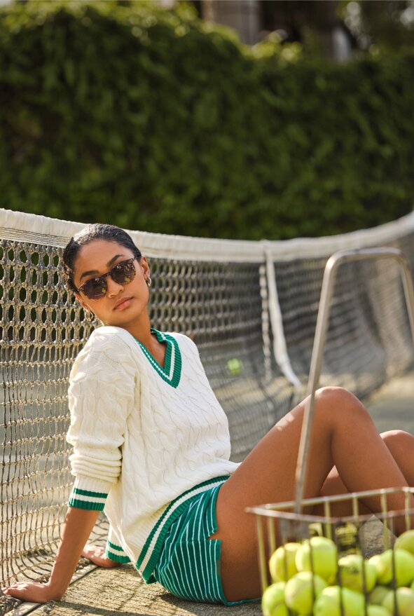 Image of a model wearing a white cable knit sweater with green striped trim, white and green striped shorts and sunglasses, sitting on a tennis court next to the net