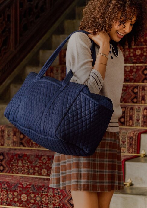 Woman carrying a blue quilted Vera Bradley duffel bag—celebrate Vera’s birthday with 25% off in store and online. Shop Vera Bradley.