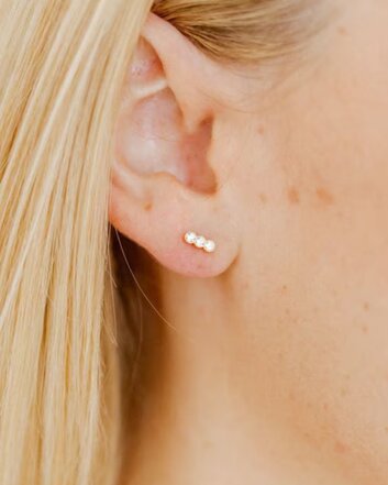 Close-up of a person with blonde hair wearing three small gemstone stud earrings in the earlobe.