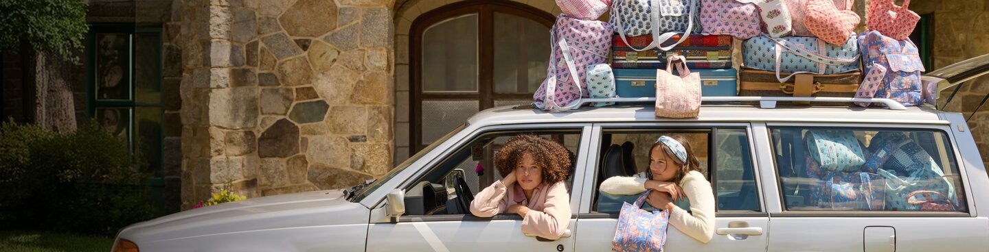 Two women lean out the windows of a white station wagon packed with colorful floral and quilted Strawflower bags, parked in front of a stone building.