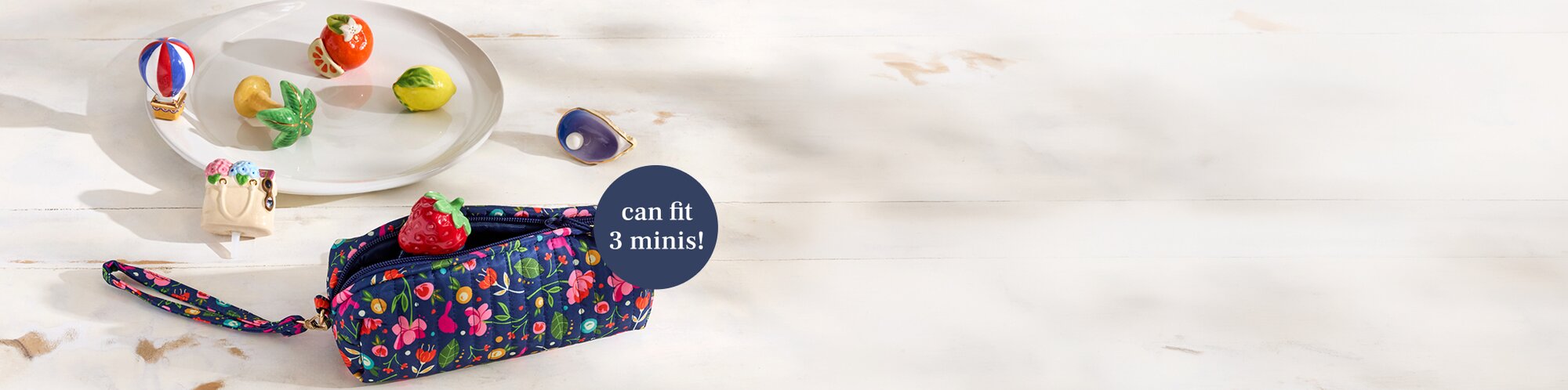 A navy floral zippered Nora Fleming pouch with a wrist strap sits on a light wooden surface, surrounded by small colorful Mini Ornaments like lemon, orange, strawberry,  hot air balloon, with a label indicating it can fit three minis.