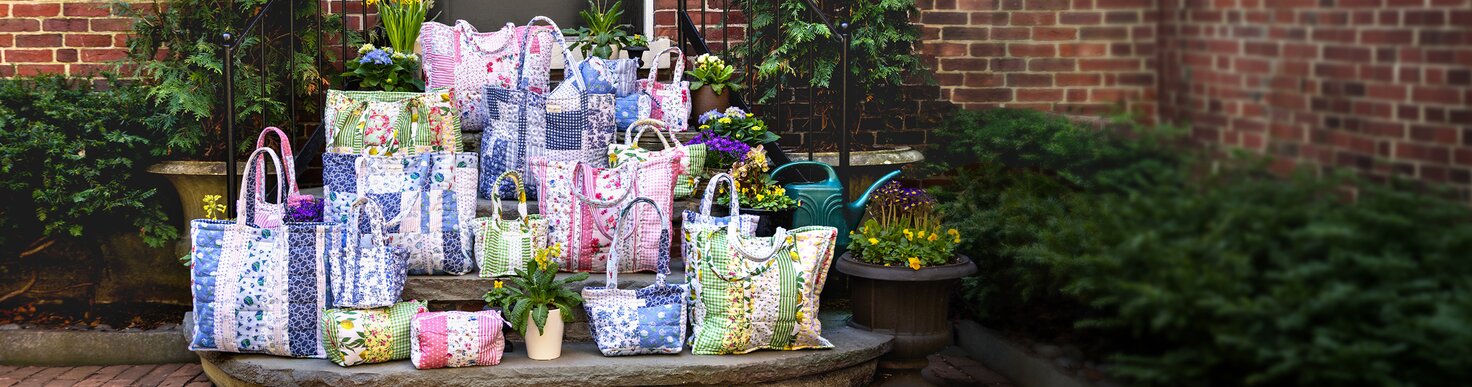 A woman sits on an outdoor tennis court in warm sunlight, wearing sunglasses, a light jacket, and sneakers. Beside her are colorful Stawflower bags, including a tote holding a tennis racket and a duffel holding gear, styled for a sporty, casual day.