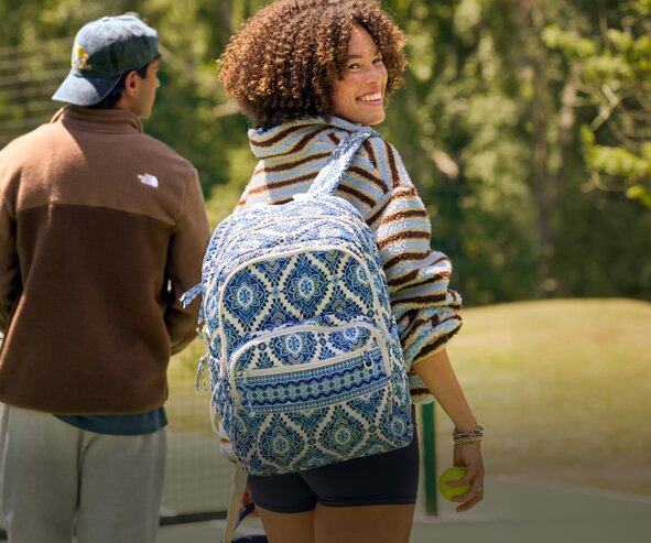 A smiling girl wearing a blue patterned Vera Bradley backpack looks back over their shoulder while walking outdoors with another person on a sunny day. They hold a tennis ball, with trees and a tennis court in the background.