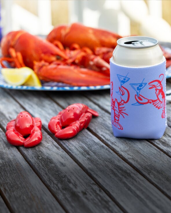 Blue can cooler with red lobster illustrations on a weathered picnic table beside lobster-themed salt & pepper shakers and seafood.