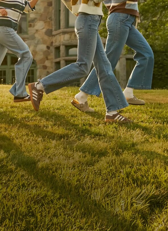 Three people walk across a sunny campus lawn wearing denim jeans and cozy knit layers, with a stone academic building in the background.