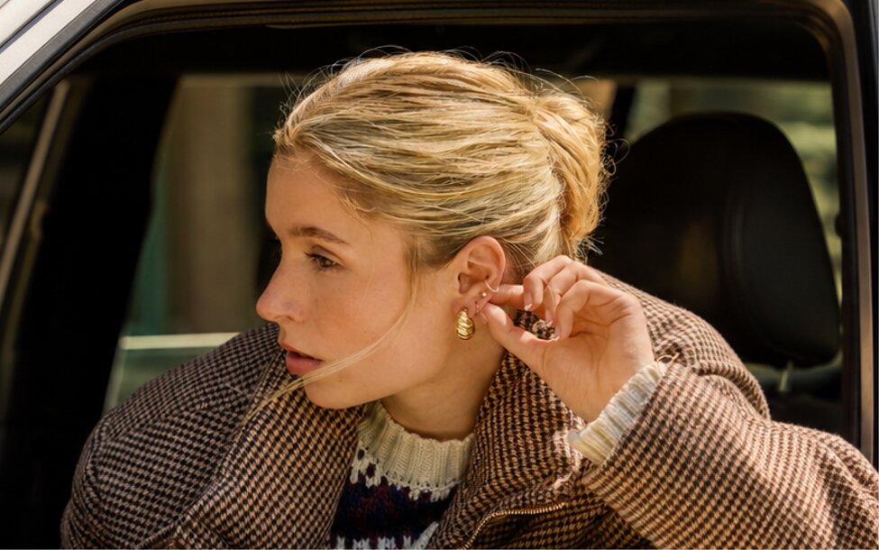 Person putting on gold earrings while seated in a car. Shop jewelry.