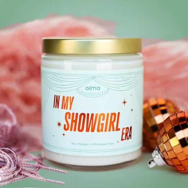 Glass candle labeled "IN MY SHOWGIRL ERA" with a gold lid and light blue label 