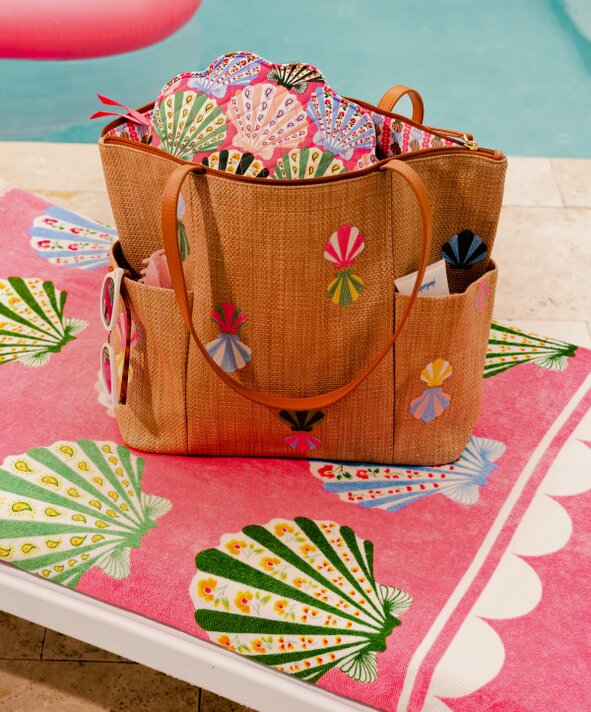 Vera Bradley faux straw beach tote with colorful embroidered shell details sitting on a Vera Bradley pink shell patterned beach towel beside a pool. The open tote is filled with matching printed accessories, with sunglasses tucked into the side pocket and a bright, resort-style poolside setting in the background.