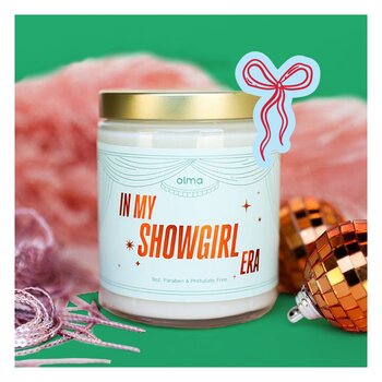 Glass candle labeled "IN MY SHOWGIRL ERA" with a gold lid and light blue label 