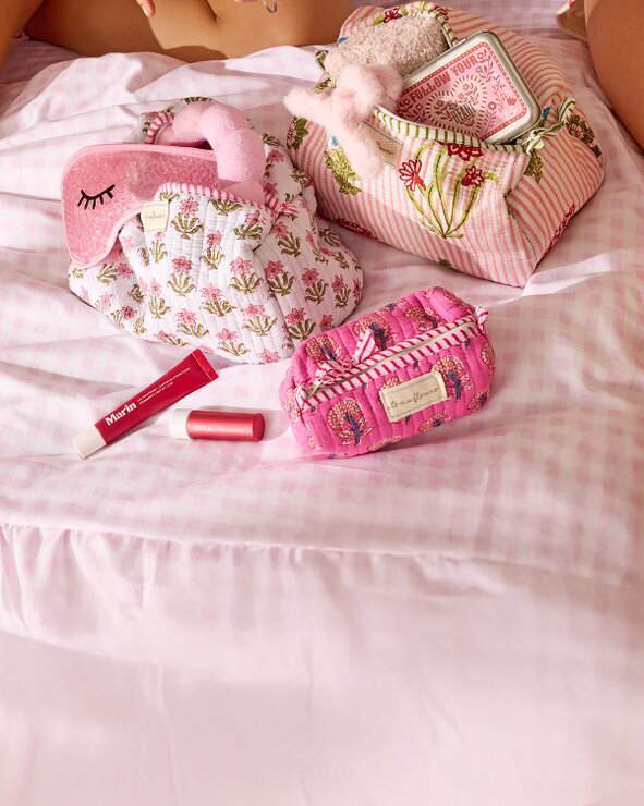 Image of strawflower cosmetic bags on a bed, filled with Marin lip balm, an eye mask, headband, Boy Smells body spray, hair clips, Touchland