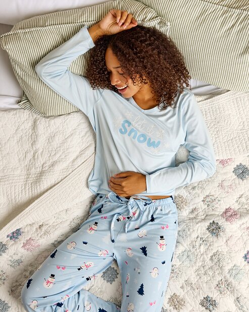 Woman lounging on a bed in light blue snowman-print pajamas with a "Let It Snow" top.