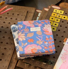 Floral-print Strawflower journals stacked on vintage pattern-making boards with colored pencils nearby.