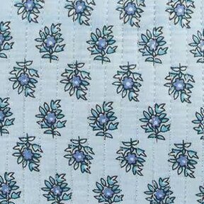 Close-up of a quilted fabric with a repeating small floral motif in blue and navy on a pale blue background. Vertical stitched lines run through the fabric, and a narrow blue-and-white striped binding borders the top edge.