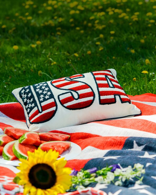 Patriotic picnic setup with an American flag blanket, strawberries, watermelon, flowers, and woven picnic basket spread across green grass.