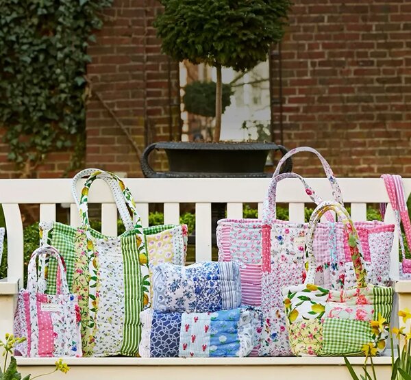 Colorful handmade quilted Strawflower tote bags and cushions in floral and patchwork patterns displayed outdoors on a bench, surrounded by potted flowers and a watering can.