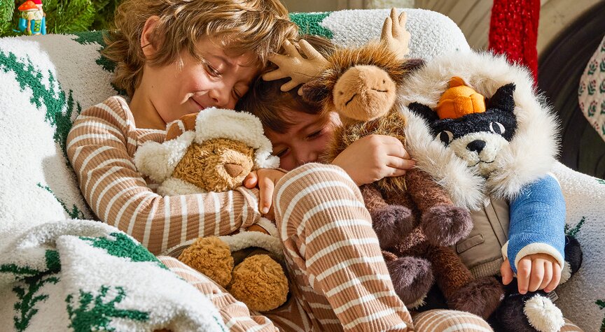 Two children in matching striped pajamas cuddle on a cozy couch with Jellycat plush toys, surrounded by festive holiday blankets.