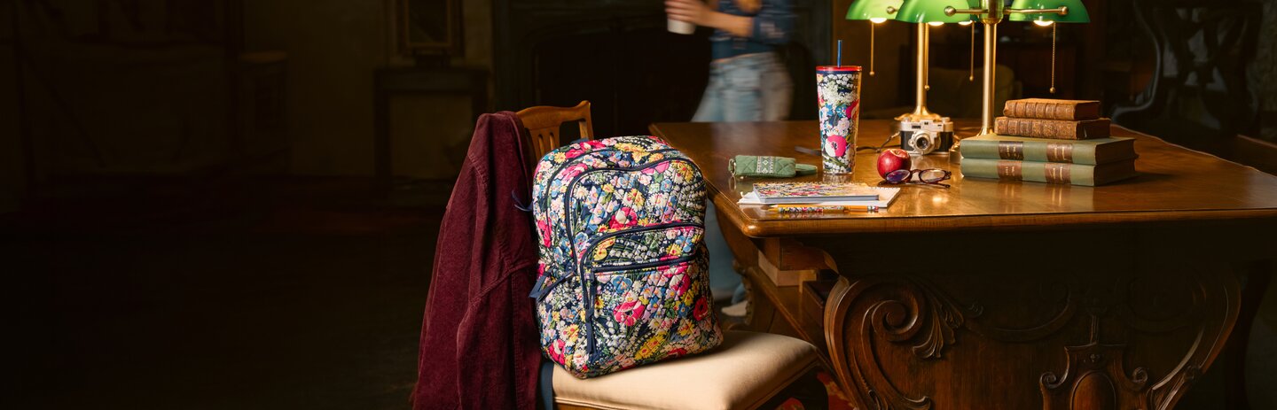 Floral Vera Bradley backpack and school essentials on a library table—get 25% off in store and online. Shop Vera Bradley.