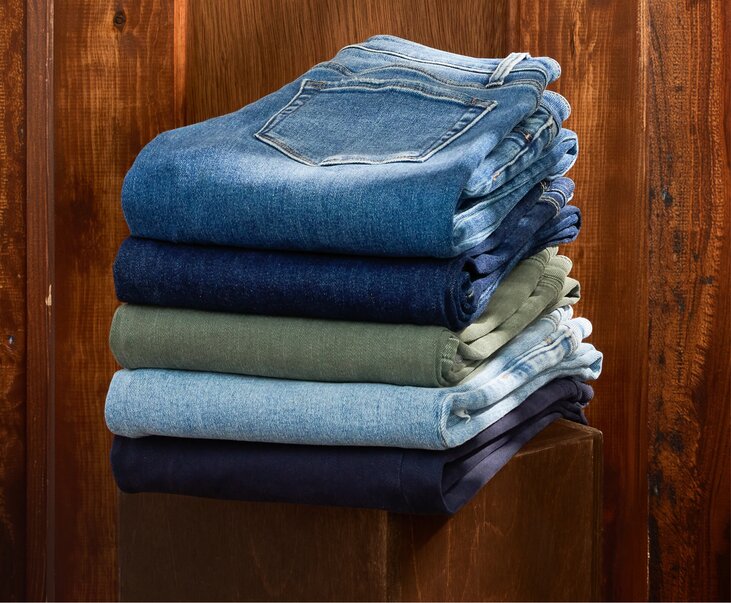 A stack of folded denim jeans in light, medium, and dark washes is displayed on a wooden shelf against a wood-paneled backdrop.