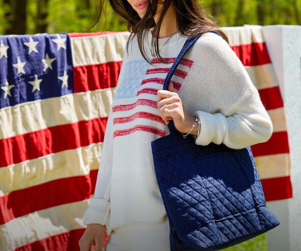 Person wearing a cream American flag sweater and carrying a navy Vera Bradley  quilted tote bag in front of a hanging American flag outdoors.