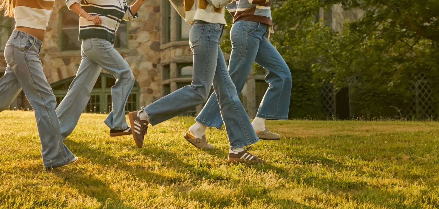 Three people walk across a sunny campus lawn wearing denim jeans and cozy knit layers, with a stone academic building in the background.