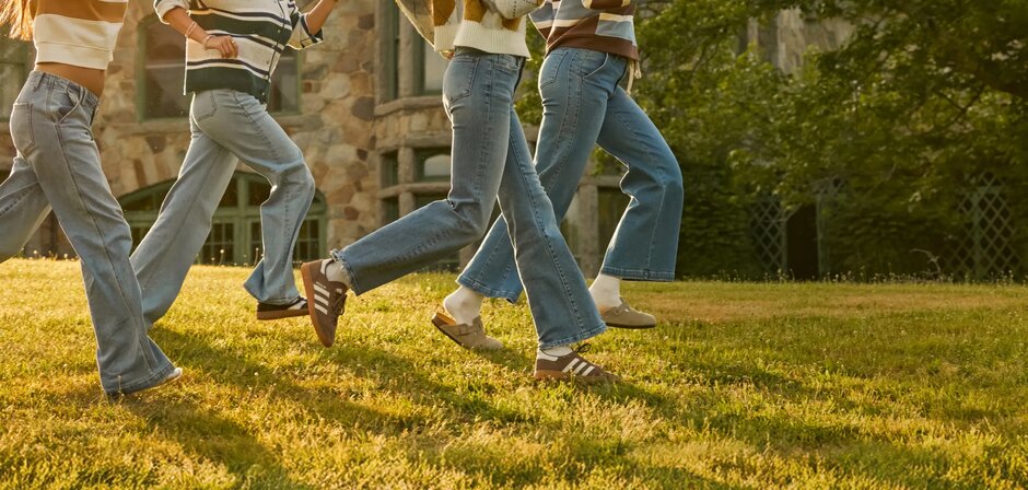 Three people walk across a sunny campus lawn wearing denim jeans and cozy knit layers, with a stone academic building in the background.