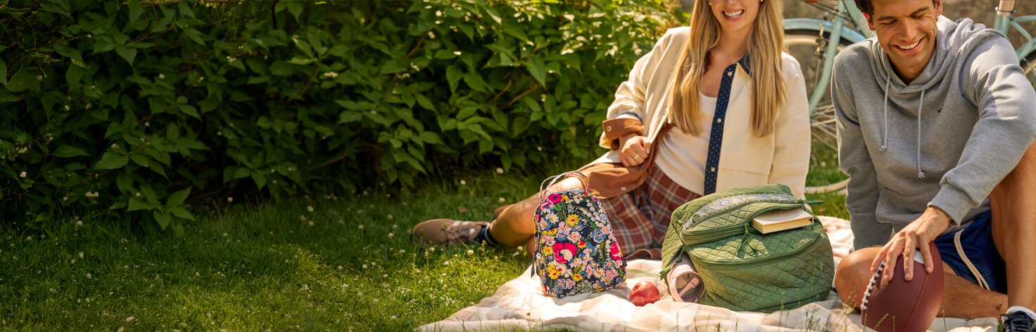 Two people enjoying a picnic on a blanket in a grassy park with a colorful Vera Bradley backpack and lunchbag, a football, books, and an apple nearby. Bicycles lean in the background, creating a relaxed back-to-school scene.