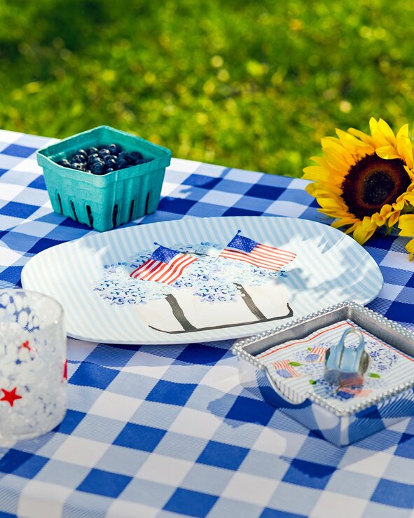 Patriotic outdoor picnic table setting featuring blue-and-white gingham linens, decorative serving pieces, fresh blueberries, and sunflower accents.