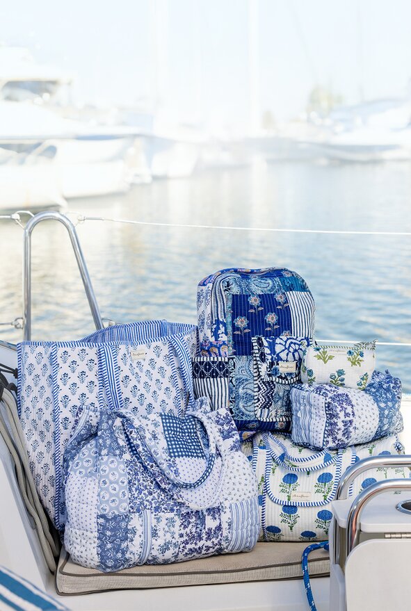 Three overlapping instant photos placed on folded blue-and-white striped and denim fabrics. The center photo shows a smiling woman sitting on a sailboat in a navy top and light blue sailboat-print jeans, shading her eyes from the sun. The left photo shows a cropped view of someone in a navy sweater and flared jeans standing barefoot on a dock, and the right photo shows a person in a pink sweater and jeans sitting on a boat seat by the water.