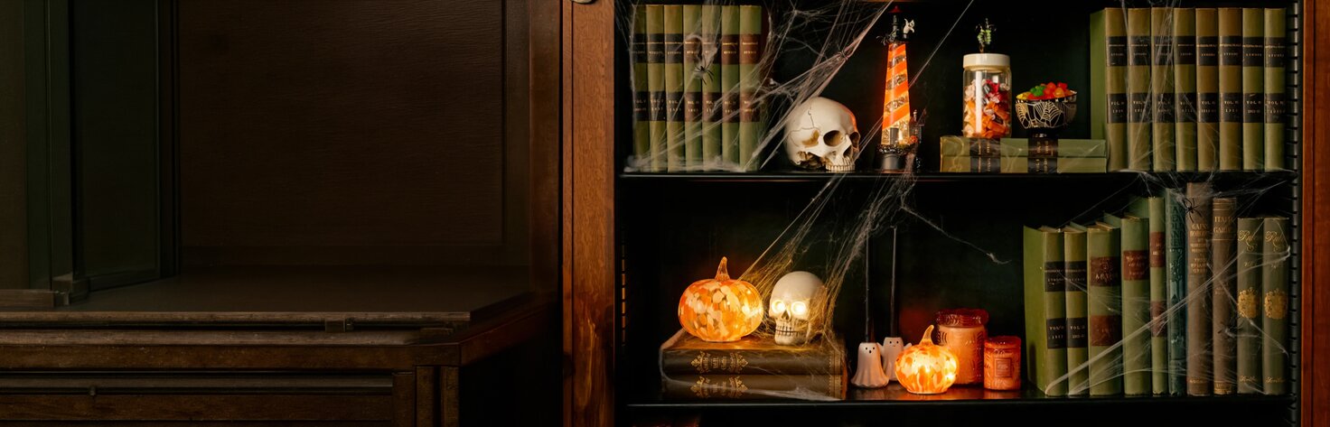 A bookshelf decorated with glowing pumpkins, skull accents, candles, cobwebs, and vintage books creates a cozy Halloween-inspired home décor display.