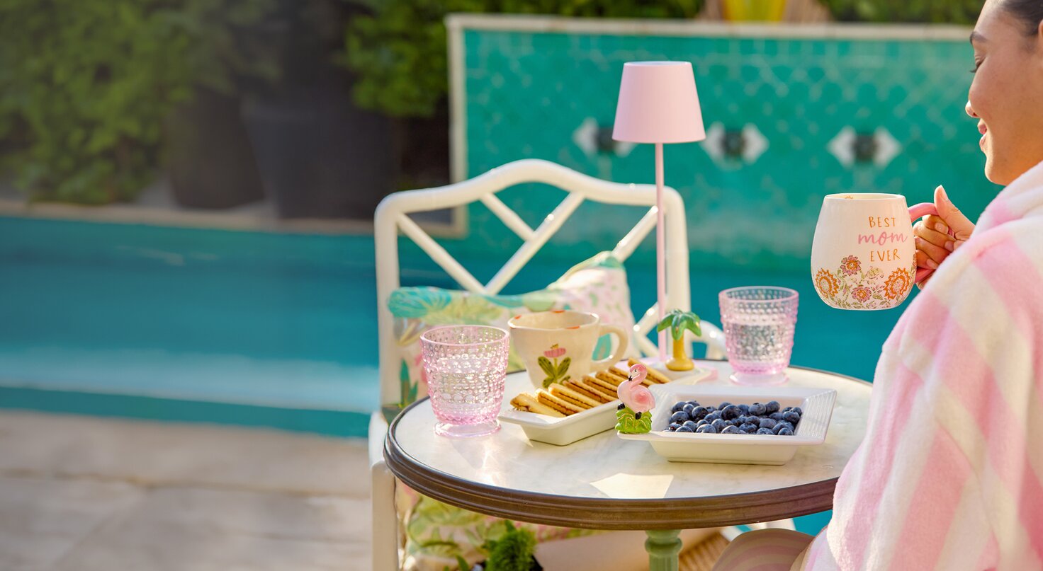 A woman in a pink and white striped robe sits by a pool, holding a Strawflower mug that says “Best Mom Ever,” next to a small table set with Nora Fleming trays holding blueberries and cookies, and decorative glasses in a sunny poolside setting.