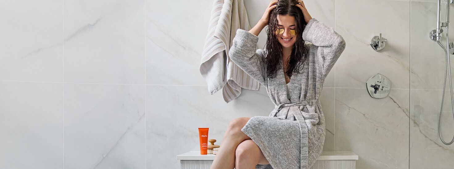 Model in a bathroom wearing a gray robe and under eye masks, with Marin skincare next to her