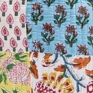 Close-up of a quilted patchwork fabric with vertical panels of different hand-drawn patterns, including stylized flowers, leaves, and fruit motifs. The colors are soft and cheerful—light blue, pink, yellow, green, and cream—with visible stitched lines running vertically through the fabric.