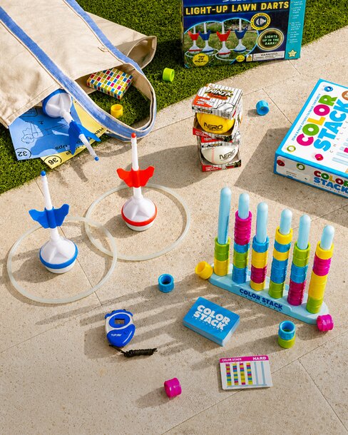 A collection of colorful outdoor games and toys is arranged on a patio beside artificial grass, including ring toss, stacking games, foam balls, and light-up lawn darts. A large canvas tote bag and vibrant packaging add to the playful backyard summer atmosphere.