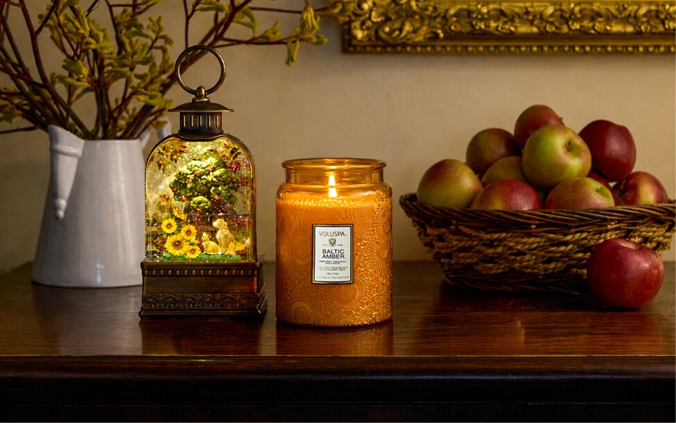 A lit amber candle and illuminated decorative lantern are displayed on a wooden table with a basket of apples and seasonal greenery in the background.