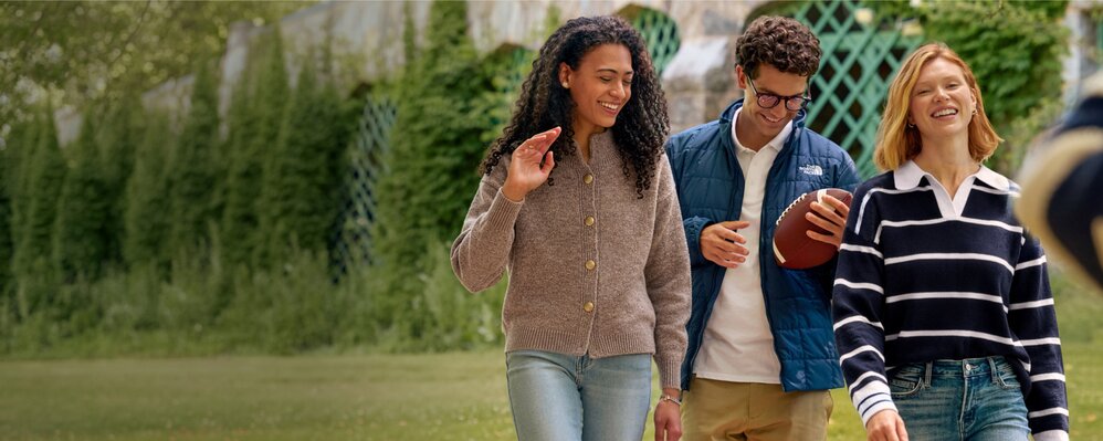 Three friends walk across a grassy lawn carrying a football, wearing casual fall clothing outdoors.