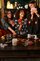 Three people gathered around a table playing cards in a book-lined room decorated with candles, seasonal treats, and Halloween-themed accents.