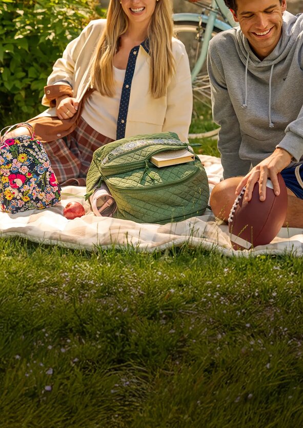 Two people enjoying a picnic on a blanket in a grassy park with a colorful Vera Bradley backpack and lunchbag, a football, books, and an apple nearby. Bicycles lean in the background, creating a relaxed back-to-school scene.