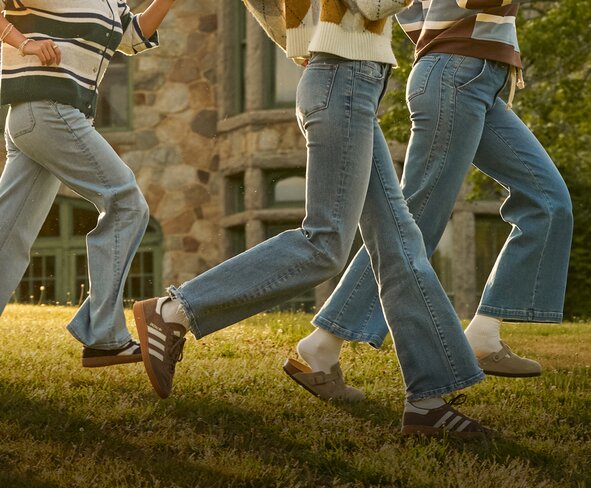 Three people jogging across a grassy lawn in front of a stone building. The image focuses on their lower bodies, showing casual jeans, sweaters, and sneakers in motion.