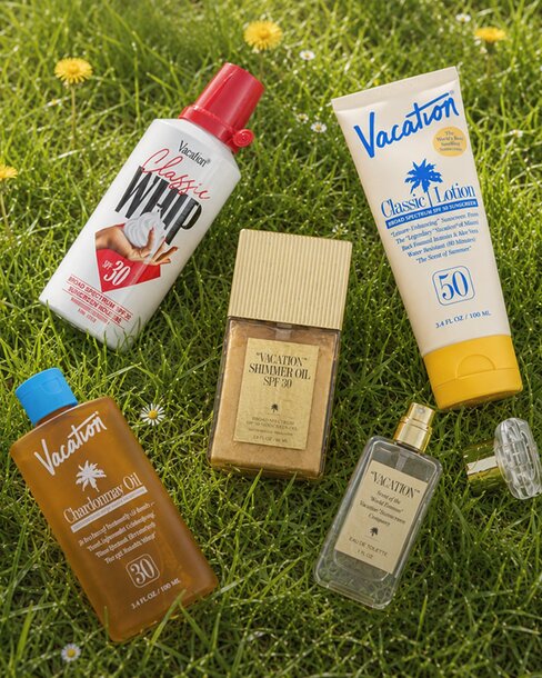 Assortment of Vacation® sunscreen, lotion, shimmer oil, body oil, and fragrance products arranged on green grass with small wildflowers.
