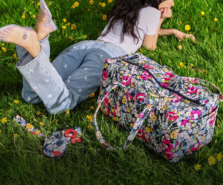 A woman lounges in the grass wearing embroidered light-wash jeans beside a colorful floral Vera Bradley duffel bag and matching pouch.