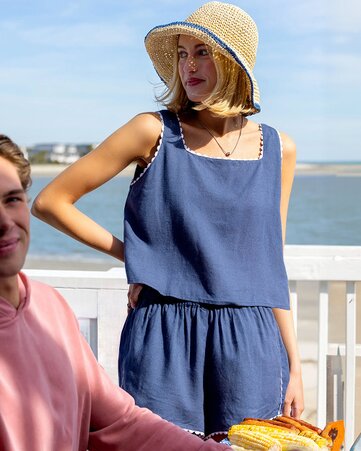 A woman wearing a navy sleeveless two-piece outfit and a woven straw sun hat stands on a waterfront deck looking toward the water. 