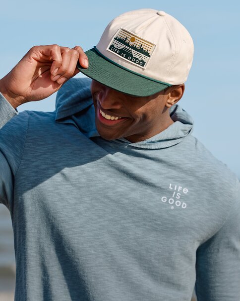 A smiling man adjusts a cream and green baseball cap with a “Life is Good” patch while wearing a light blue hooded long-sleeve shirt. The portrait is set against a clear blue sky, creating an easygoing outdoor lifestyle feel.