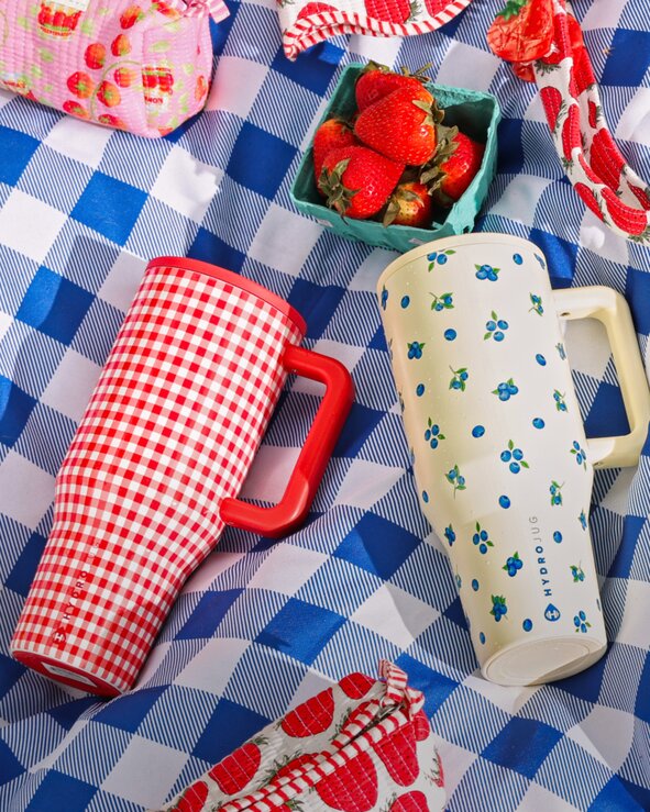 Red gingham and Blueberry patterned HydroJug tumblers, strawberry-print accessories, and fresh berries arranged on a blue gingham picnic blanket flat lay.