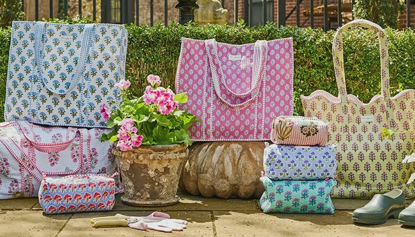 Assortment of Strawflower patchwork quilted tote bags arranged in a sunlit garden, surrounded by blooming flowers, greenery, and a pair of green garden clogs on a stone patio.