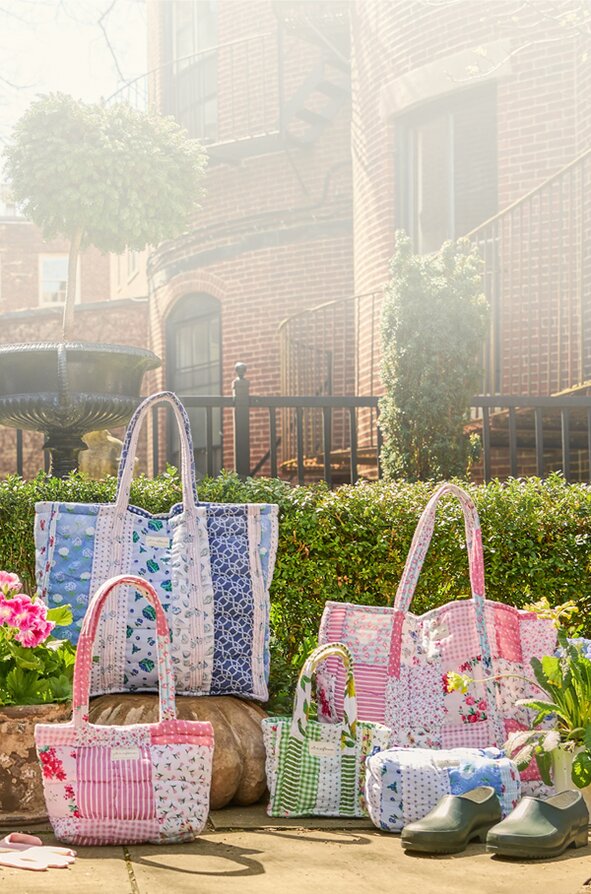 A collection of colorful, quilted patchwork Strawflower tote bags and small pouches arranged on stone steps in front of a black door and red brick house, surrounded by blooming spring flowers in pots.