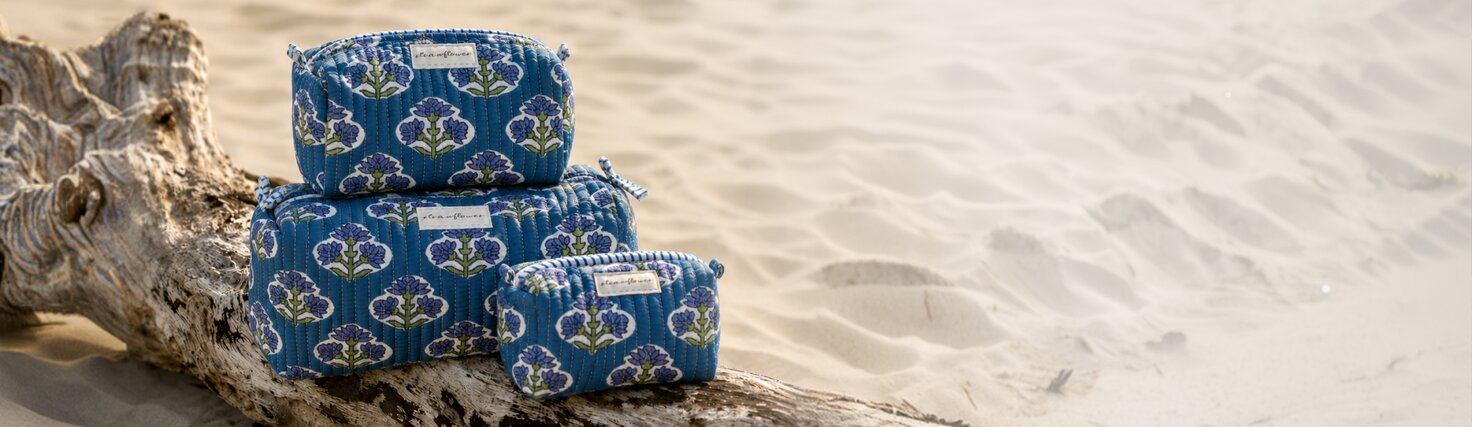 A set of three Strawflower blue floral quilted pouches displayed on driftwood in the sand.