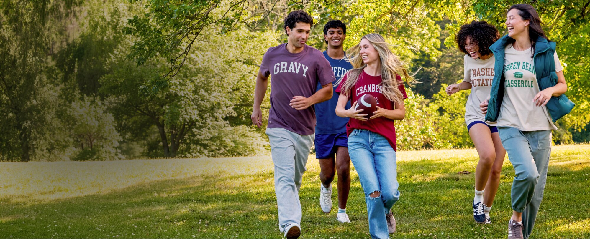 Five friends running across a grassy park in casual Thanksgiving-themed graphic tees, with one holding a football.