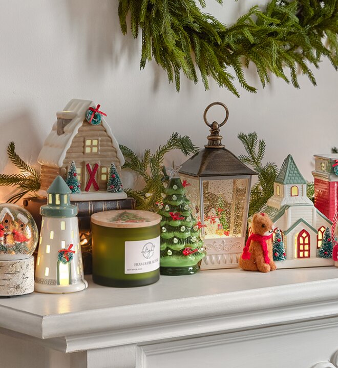 Cozy Christmas mantel decorated with illuminated ceramic houses, a snow globe, Christmas tree, lantern, candle, and festive greenery.
