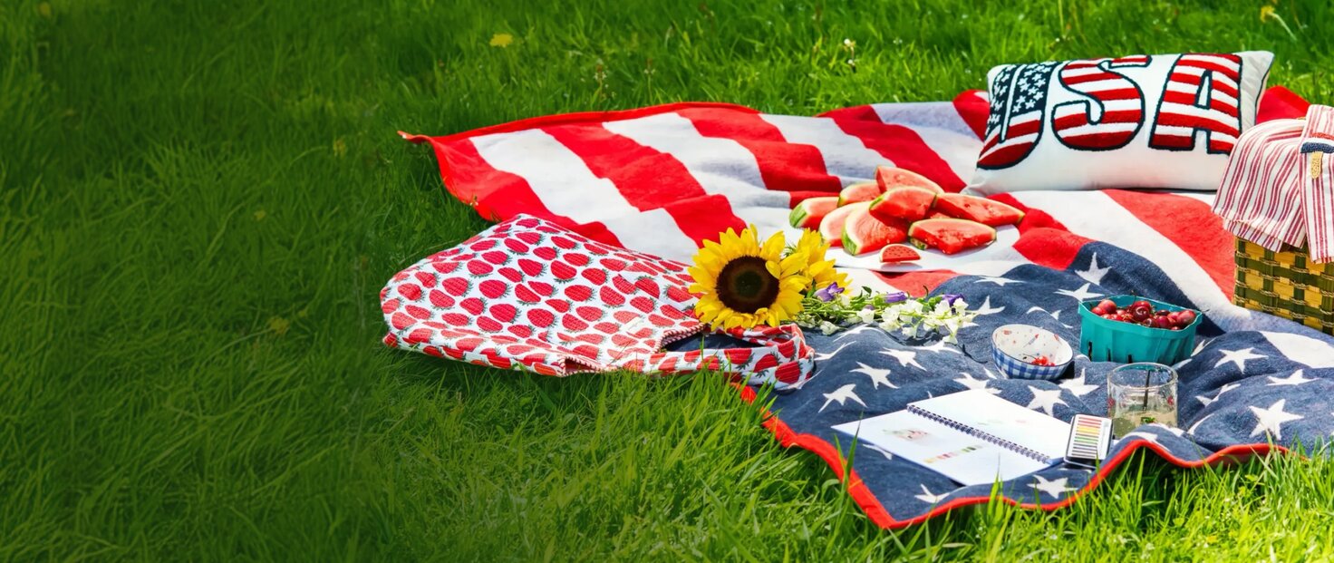 Patriotic picnic setup with an American flag blanket, strawberries, watermelon, flowers, and woven picnic basket spread across green grass.